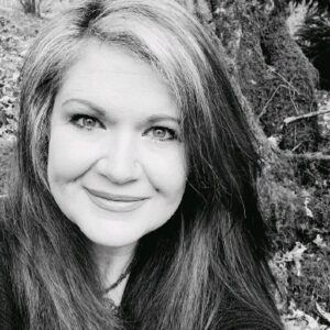 Black and white headshot of Anita smiling with long hair, wearing a dark colored shirt and sitting outside in a forest area. 