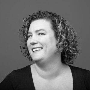 Black and white headshot of Julian smiling with her head turned to the side. She has shoulder length curly hair and is wearing a black shirt.